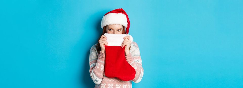 Cute girl cover face with christmas stocking, staring right with cunning gaze, standing in Santa hat and celebrating winter holidays, blue background.