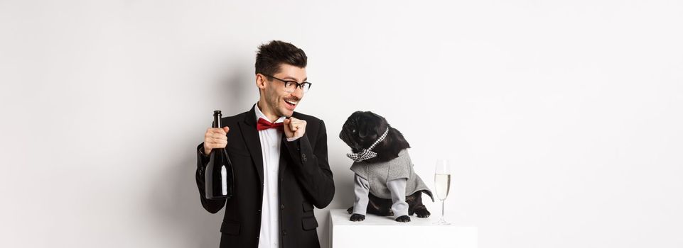 Cheerful young man in suit celebrating New Year with pet, dog and guy looking at each other, owner drinking champagne, standing over white background.