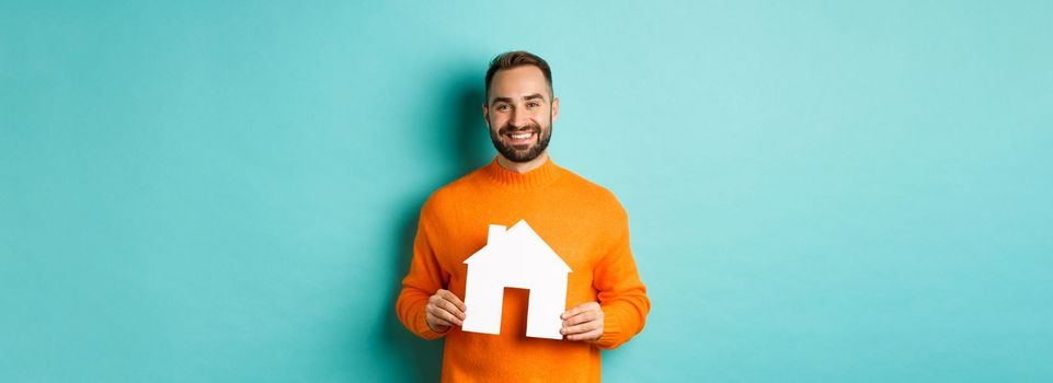 Real estate concept. Happy young man searching for home rent, holding house paper maket and smiling, standing over blue background.