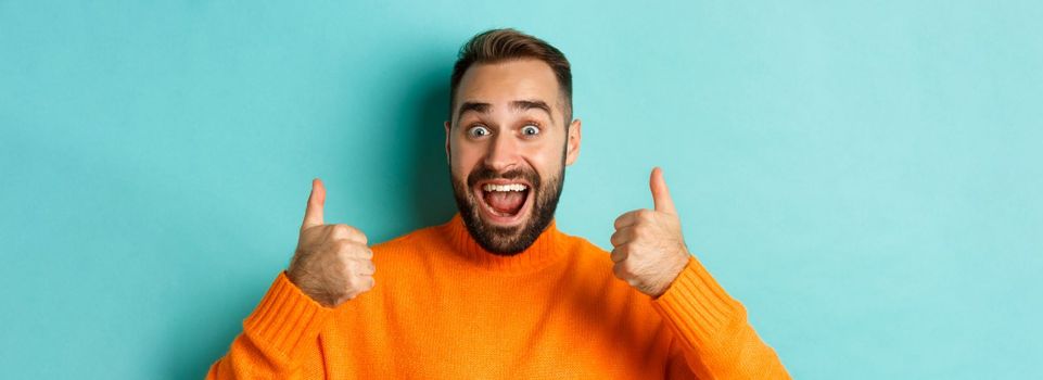 Close-up of excited handsome man showing thumb-up, recommending product, praising great quality, standing over light blue background.