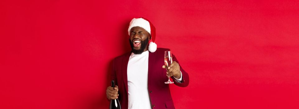 Christmas, party and holidays concept. Funny drunk Black man celebrating New Year, laughing and having fun, holding glass and bottle of champagne, red background.