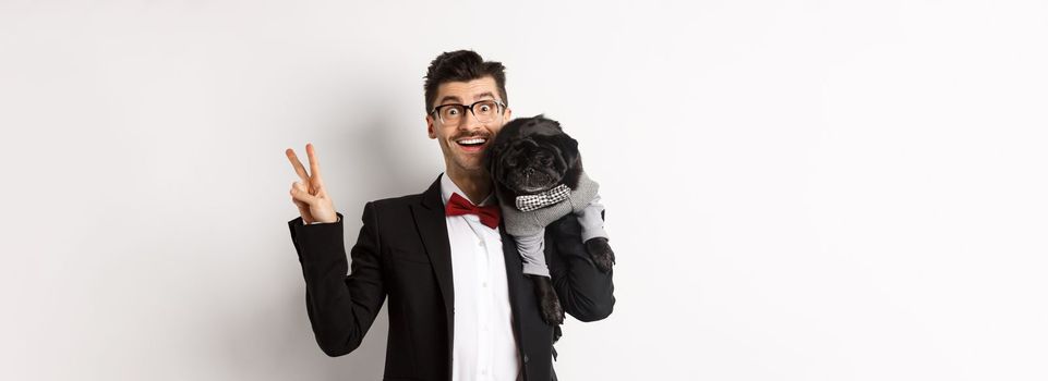 Cheerful young man in suit and glasses taking photo with cute black pug dog on his shoulder, smiling happy and showing peace sign, posing over white background.