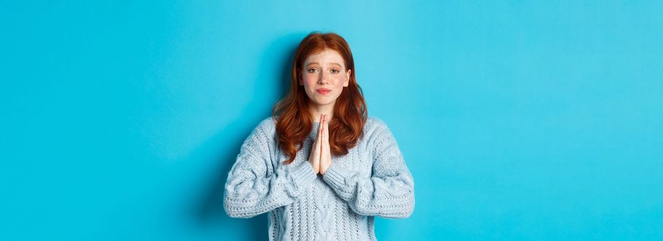 Cute teenage redhead girl asking for help, smiling while begging for favour, need something, standing over blue background.
