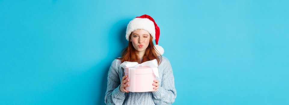 Winter holidays and Christmas Eve concept. Sassy redhead girl in sweater and Santa hat, holding New Year gift and looking at camera, standing against blue background.