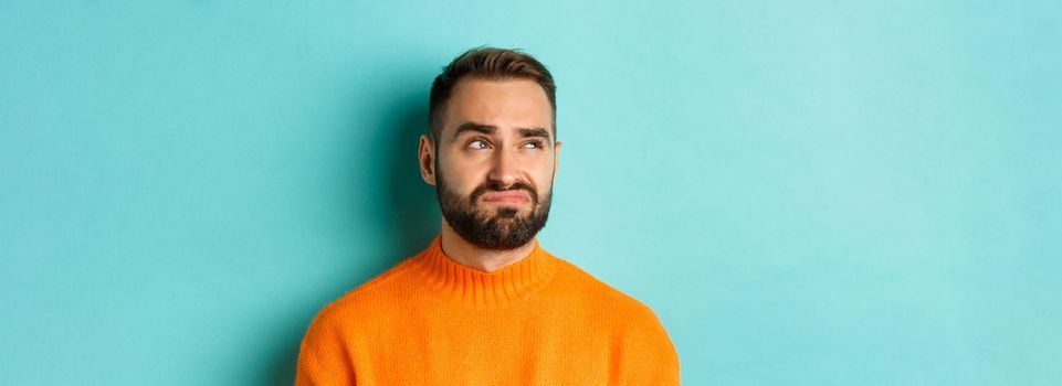 Close-up of handsome caucasian man looking left disappointed and skeptical, staring at logo, wearing orange sweater, standing against turquoise background.