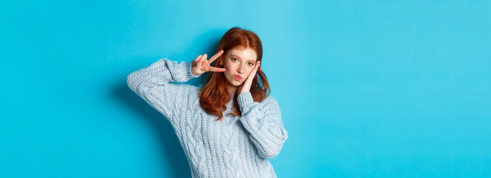 Modern teen girl with red hair, showing peace sign and posing in sweater against blue background.