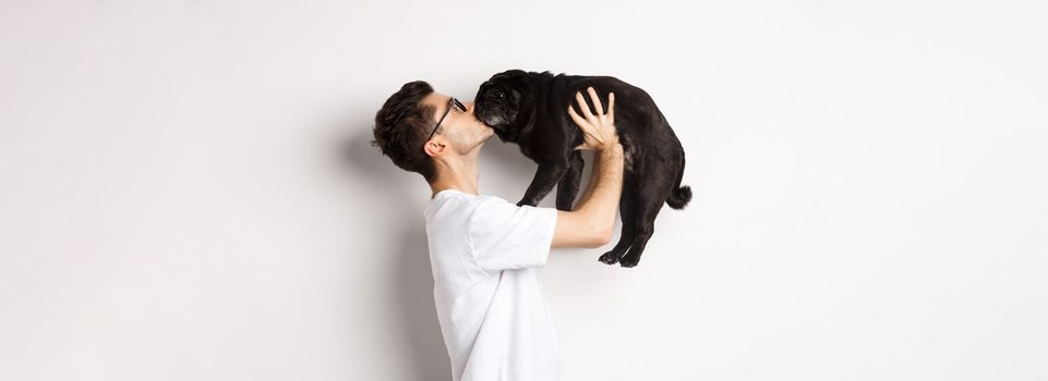 Profile of handsome young man kissing small cute dog face. Hipster guy loving his pug, standing over white background.