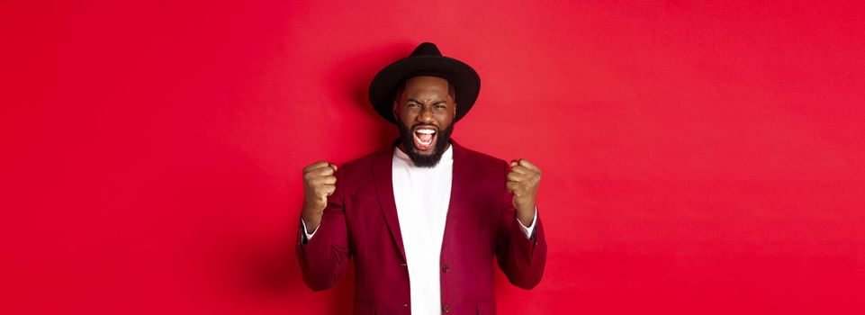 Satisfied lucky Black man celebrating victory, screaming from rejoice, winning prize, saying yes and fist pump, red background. Copy space