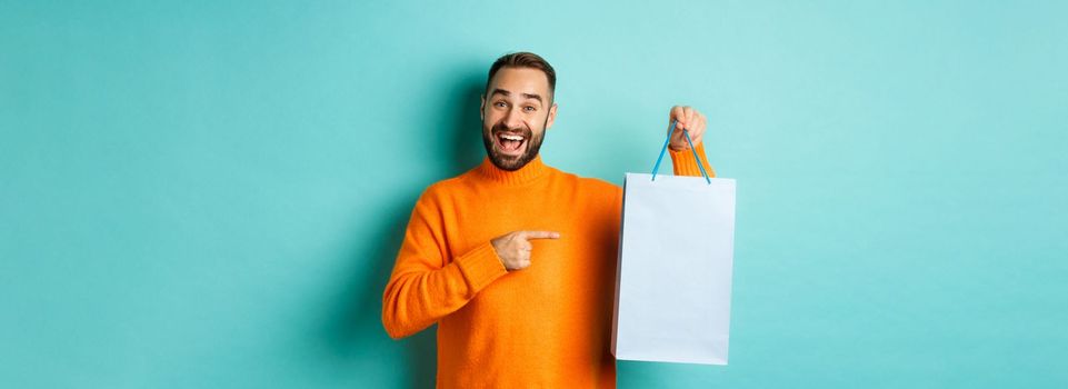 Handsome smiling man pointing finger at shopping bag, buying in stores, standing satisfied over blue background.