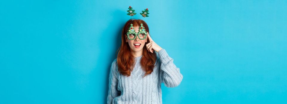 Christmas party and celebration concept. Cute redhead teen girl celebrating New Year, wearing xmas tree headband and funny glasses, looking left amused, blue background.