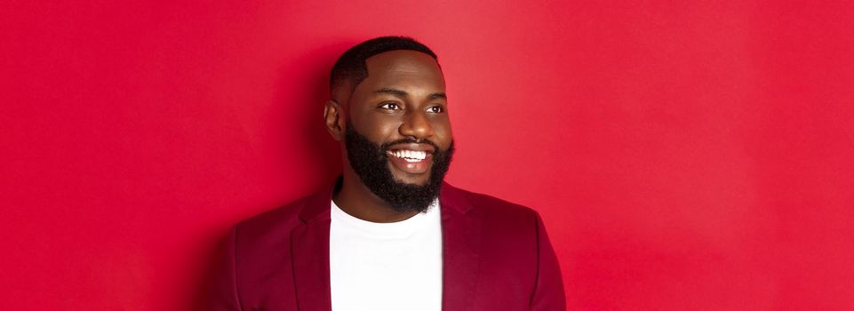 Close-up of handsome african american man with beard, looking left and smiling joyful, standing over red background.