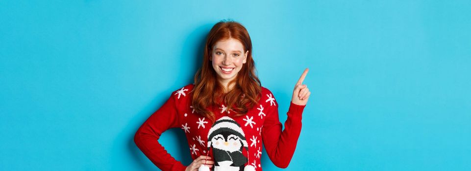 Winter holidays and Christmas Eve concept. Cute teenage girl with red wavy hair, pointing upper left corner and smiling at camera, standing over blue background.