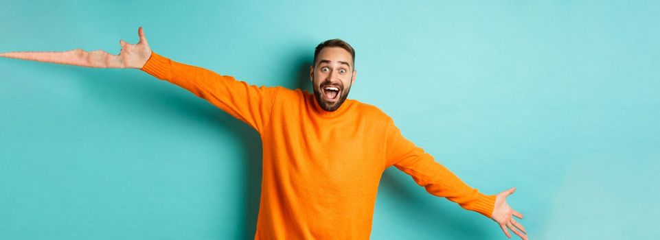 Surprised and happy man spread hands in warm welcome, reaching for hug, greeting you, showing something big, standing over light blue background.