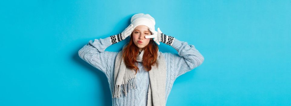 Winter and holidays concept. Happy redhead girl in beanie hat, scarf and gloves showing peace sign and pucker lips sassy, staring at camera, wearing sweater, posing over blue background.