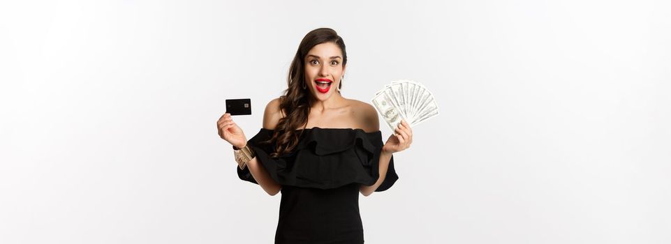 Fashion and shopping concept. Excited woman in black dress, showing credit card and dollars, smiling and staring at camera, white background.