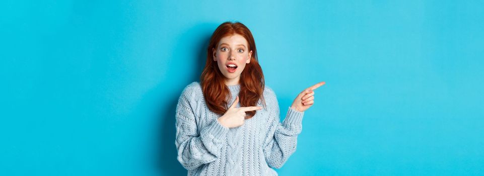 Amazed redhead girl in sweater, pointing fingers left at logo and staring excited at camera, standing against blue background.