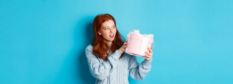 Curious teen girl with red hair, shaking gift box and wonder what inside, standing over blue background.