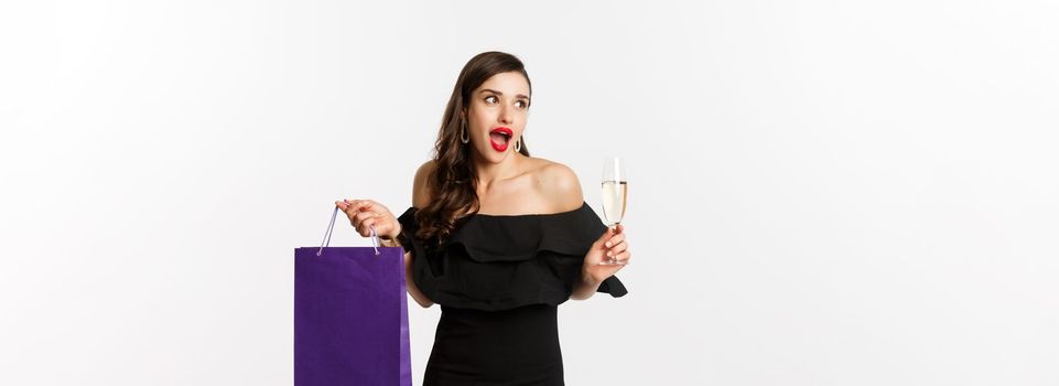 Excited woman shop and drink champagne, holding shopping bag, looking amazed, standing against white background.