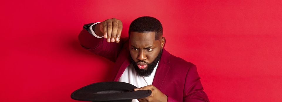 Close-up of charismatic Black male magician perform a trick with his hat, performing on christmas party, standing over red background.