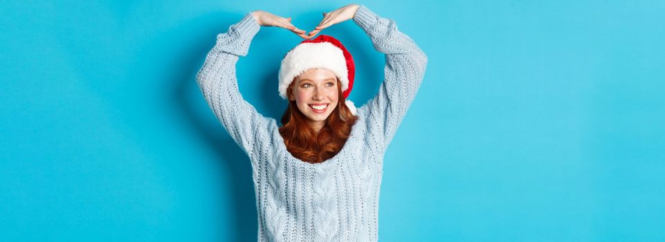 Winter holidays and Christmas Eve concept. Cute redhead teen girl in santa hat and sweater, making heart sign and smiling, wishing merry xmas, standing over blue background.