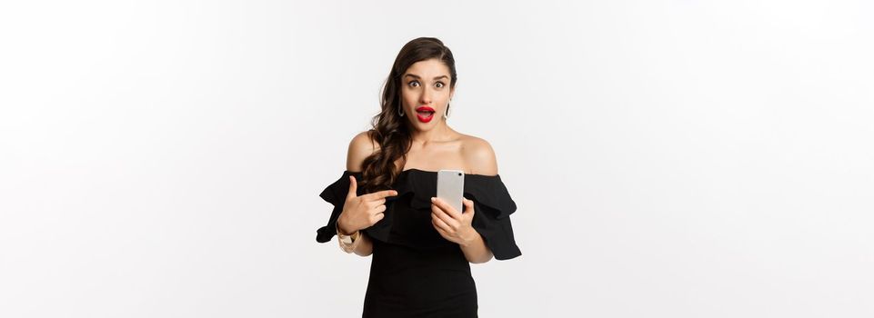 Online shopping concept. Stylish woman in black dress, wearing makeup, pointing finger at mobile phone with surprised emotion, standing over white background.