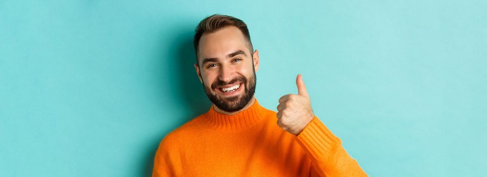 Close-up of attractive young man showing thumb-up, praising good work, standing over light blue background, giving positive answer.