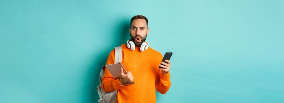 Handsome man student with headphones and backpack, holding digital tablet and smartphone, staring confused at camera, standing against turquoise background.