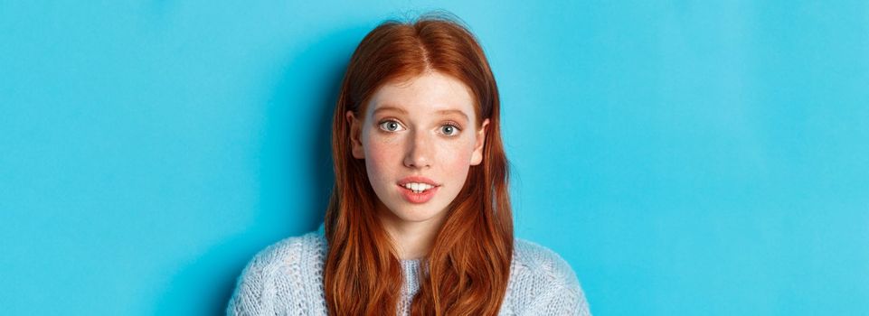 Headshot of cute redhead girl with freckles, looking hopeful and innocent at camera, standing over blue background.