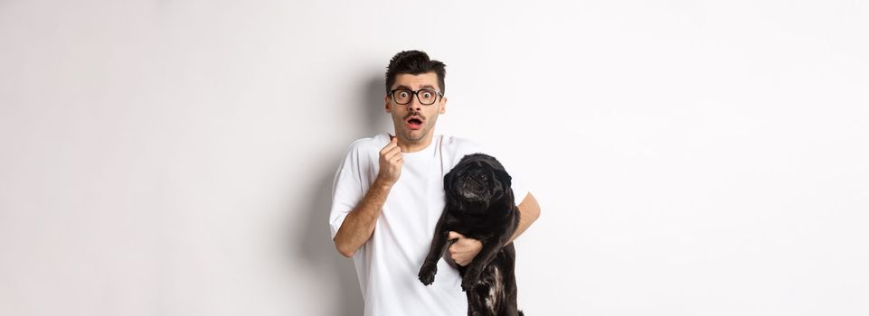 Scared hipster hugging his cute black puppy and staring at camera frightened. Dog owner looking shocked, holding pug, standing over white background.