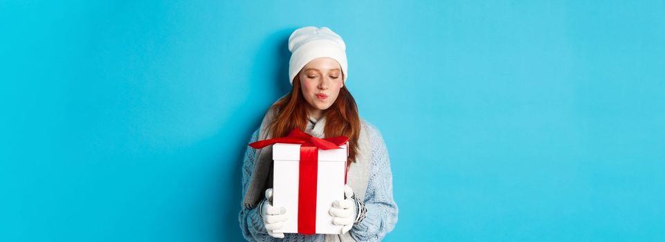 Winter holidays and Christmas sales concept. Intrigued redhead girl holding present, curiously staring at box with gift, trying guess what inside, standing over blue background.