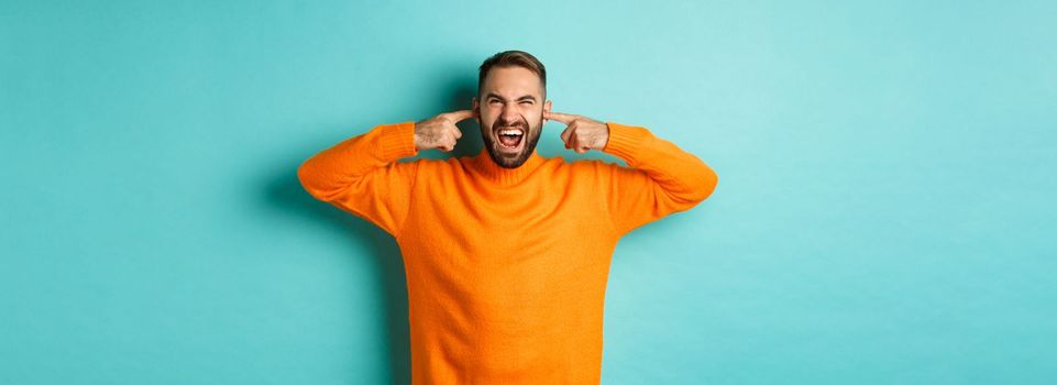 Displeased man shut ears and grimacing, loud neighbours, standing annoyed with noise, shouting angry and distressed, light blue background.