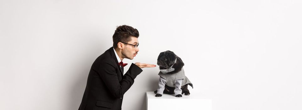 Pets, winter holidays and celebration concept. Handsome young man sending air kiss at cute black puppy wearing costume for New Year, owner standing in suit over white background.