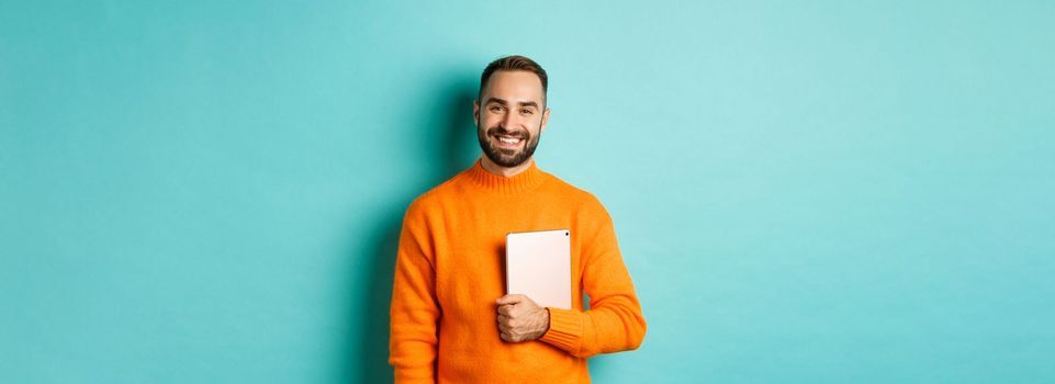 Handsome freelancer man holding laptop and smiling, standing happy over light blue background.