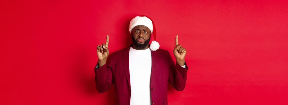 Christmas, party and holidays concept. Miserable and sad african american man pointing fingers up, looking disappointed, wearing santa hat, red background.