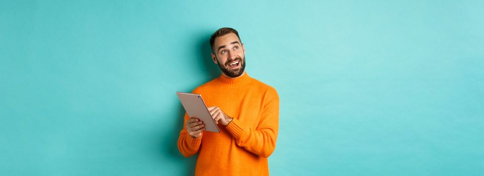 Handsome man thinking and using digital tablet, shopping online and pondering ideas, looking at upper left corner, standing over light blue background.