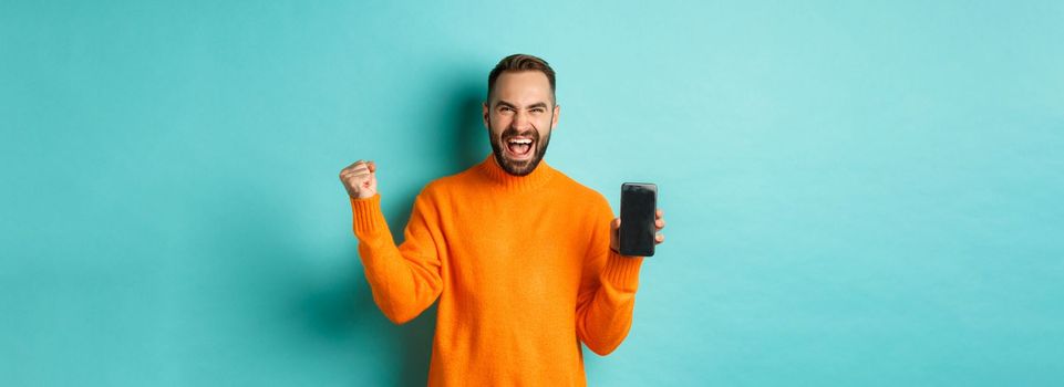 Image of happy young man winning and showing mobile phone screen, rejoicing and celebrating victory, fist pump with satisfaction, standing over light blue background.