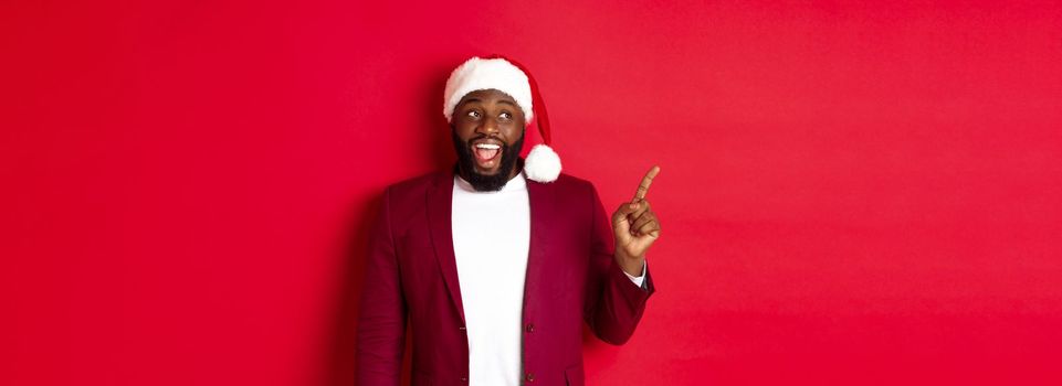 Christmas, party and holidays concept. Cheerful Black guy in santa hat pointing fingers left, showing banner, standing amused against red background.