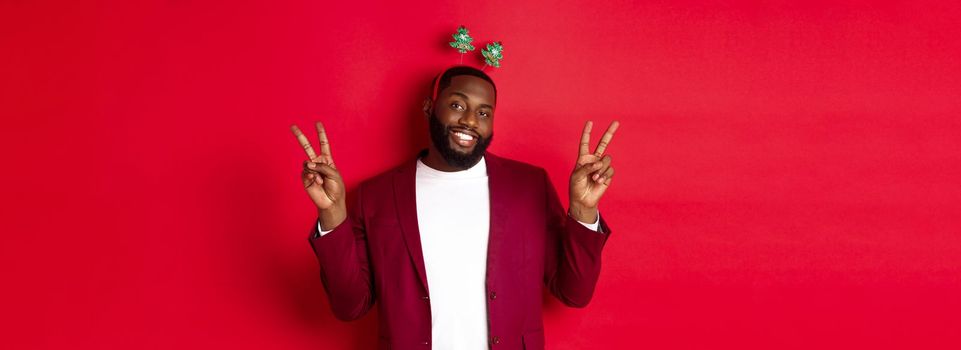 Merry Christmas. Cheerful african american bearded guy in party headband, smiling and showing peace signs, standing over red background.