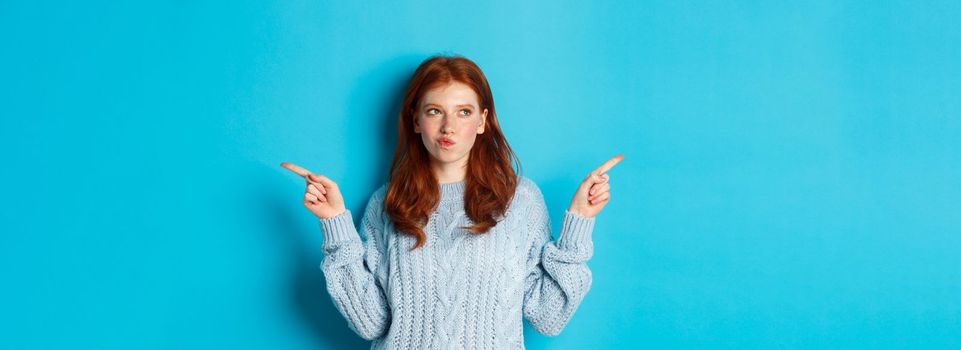 Winter holidays and people concept. Thoughtful redhead girl making decision, pointing fingers sideways, choosing between two ways, standing over blue background.
