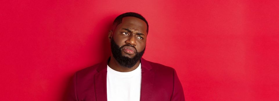 Close-up of disappointed and skeptical Black man looking left, grimacing and sulking displeased, standing over red background. Negative emotion