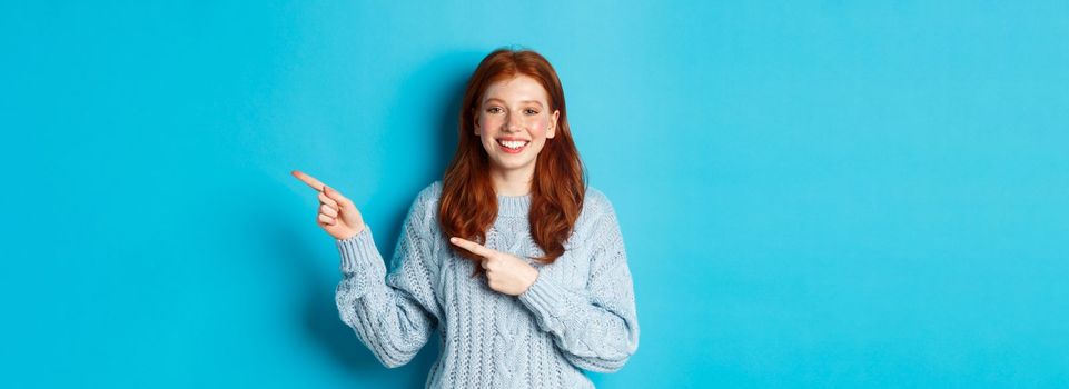 Winter holidays and people concept. Pretty teenage girl with red hair, pointing fingers right at logo copy space, smiling at camera, blue background.