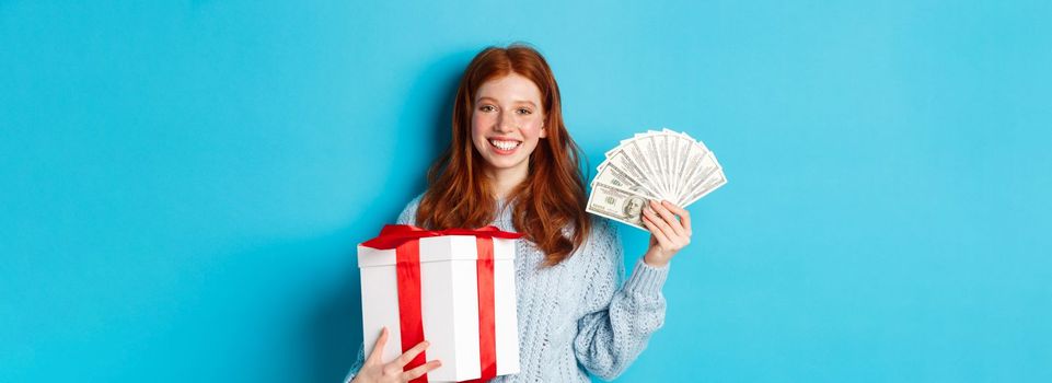 Young redhead woman holding Christmas gift box and money, smiling pleased, standing over blue background.