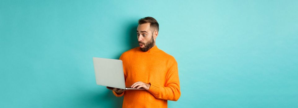 Freelance and technology concept. Image of handsome adult man staring at laptop with amazement, shopping online or working remote, standing over turquoise background.