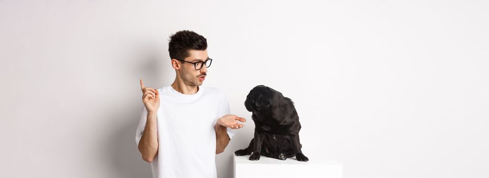 Handsome young man teaching dog commands, talking to cute black pug, standing over white background.