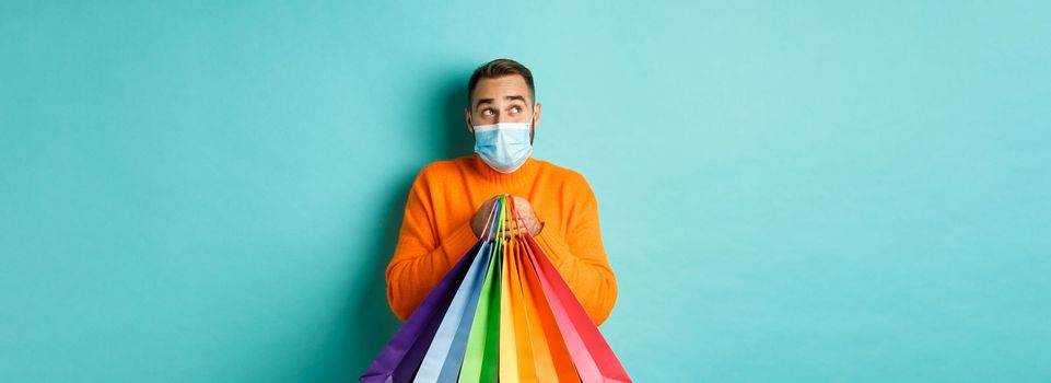 Covid-19, pandemic and lifestyle concept. Man thinking, wearing face mask, holding shopping bags, imaging something, standing over turquoise background.