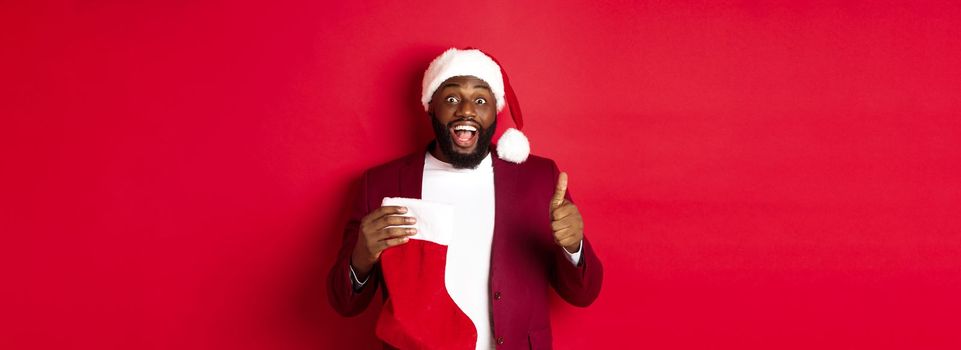 Excited Black man showing thumb-up in approval, holding christmas sock with holiday gifts, smiling amazed, standing over red background.