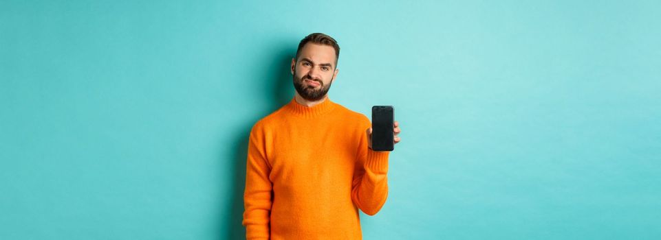 Displeased and disappointed man in orange sweater, grimacing and showing mobile screen, standing over light blue background.