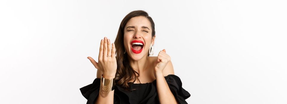 Fashion and beauty concept. Excited and happy woman said yes, showing hand with engagement ring and shouting joyful, become bride, standing over white background.