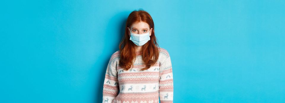 Winter, covid-19 and quarantine concept. Young redhead girl in sweater and face mask staring at camera, standing over blue background.