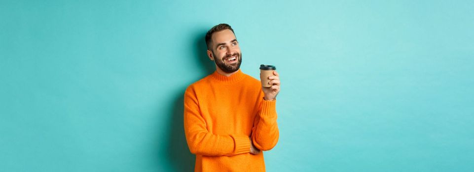 Thoughtful smiling man looking at upper left corner, having idea while drinking coffee from takeaway cup, standing in orange sweater against turquoise background.